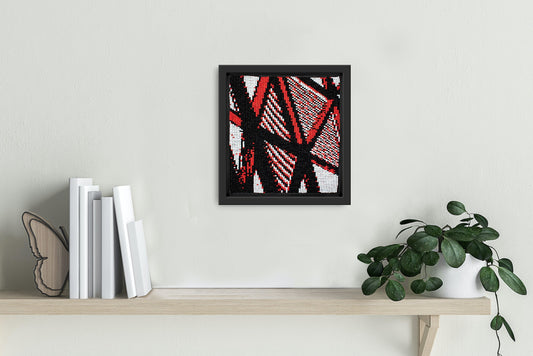 Handwoven abstract art piece with red, black, and white patterns from Gujarat hung on wall with books and plants.